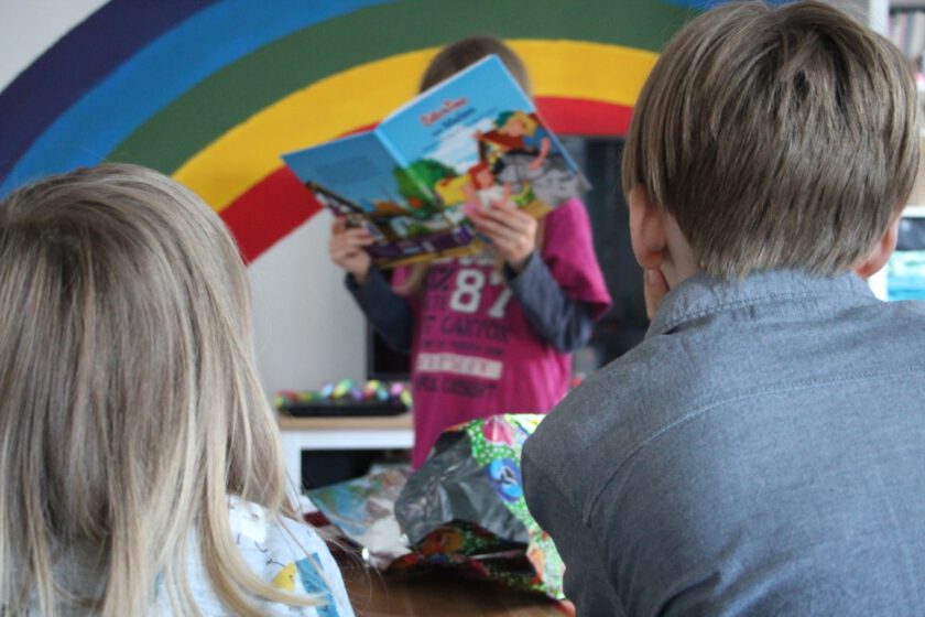 Miteinander durch die Pubertät - Inke Hummel Zwei Kinder von hinten. Vor ihnen, im Bild mittig, steht ein weiteres Kind und liest aus einem Buch vor.