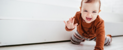 Kleinkind krabbelt auf weißem Boden und hebt dabei lächelnd eine Hand hoch. Es sieht aus als würde es eine Babygebärde zeigen.