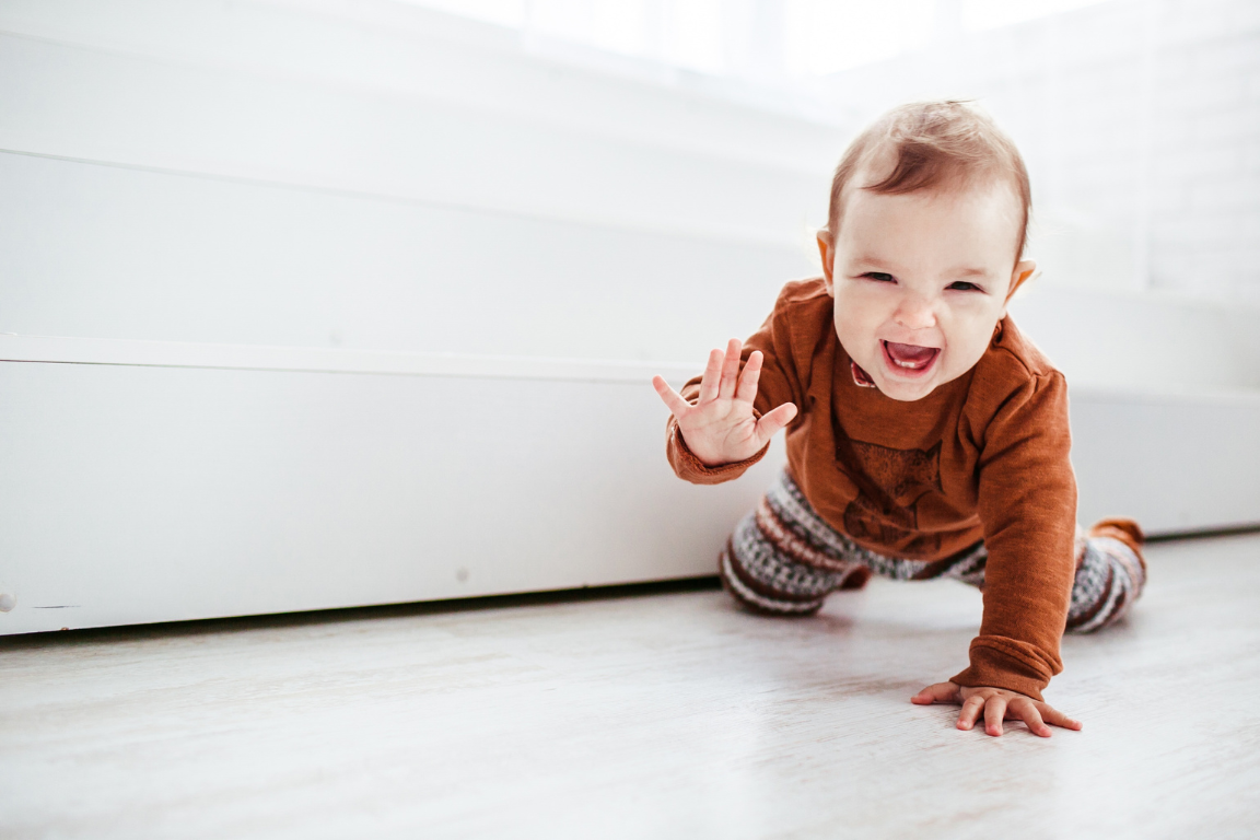 Kleinkind krabbelt auf weißem Boden und hebt dabei lächelnd eine Hand hoch. Es sieht aus als würde es eine Babygebärde zeigen.